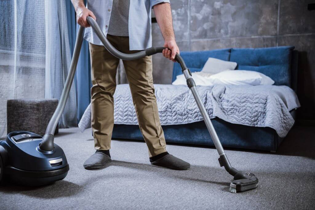 Partial view of man with vacuum cleaner cleaning carpet in bedroom